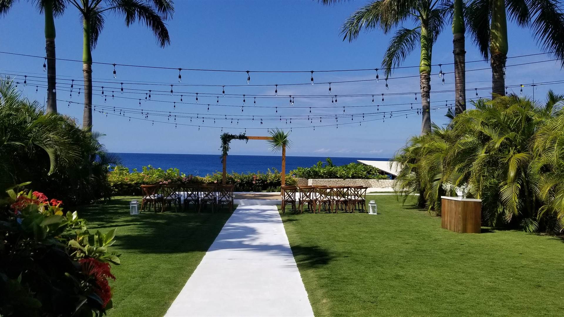A beachside wedding setup with string lights, a white aisle leading to an arch, surrounded by palm trees and lush greenery by the ocean.