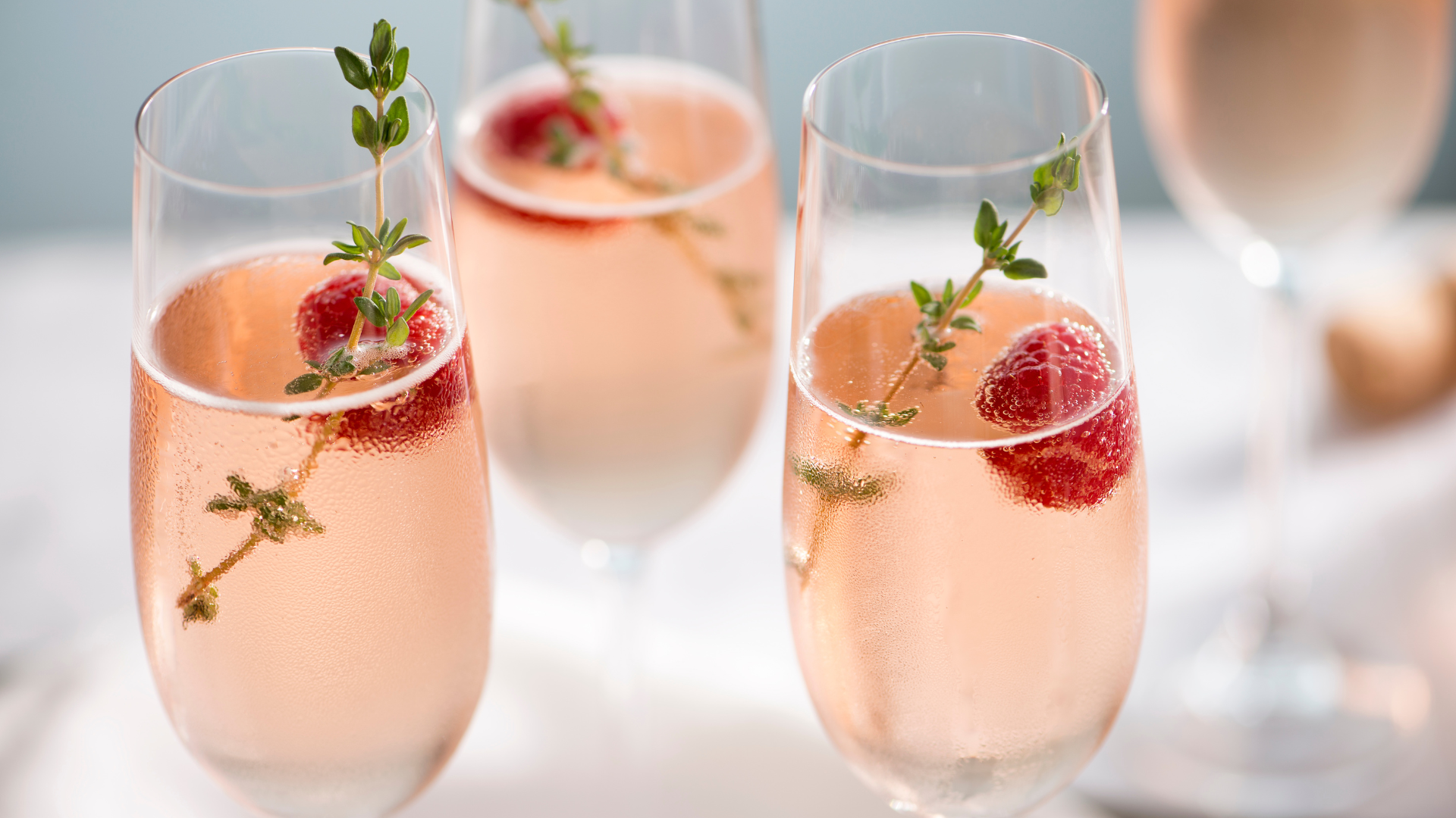 Sparkling peach rosé glasses with raspberries, thyme garnish, and delicate bubbles on a bright, festive table.