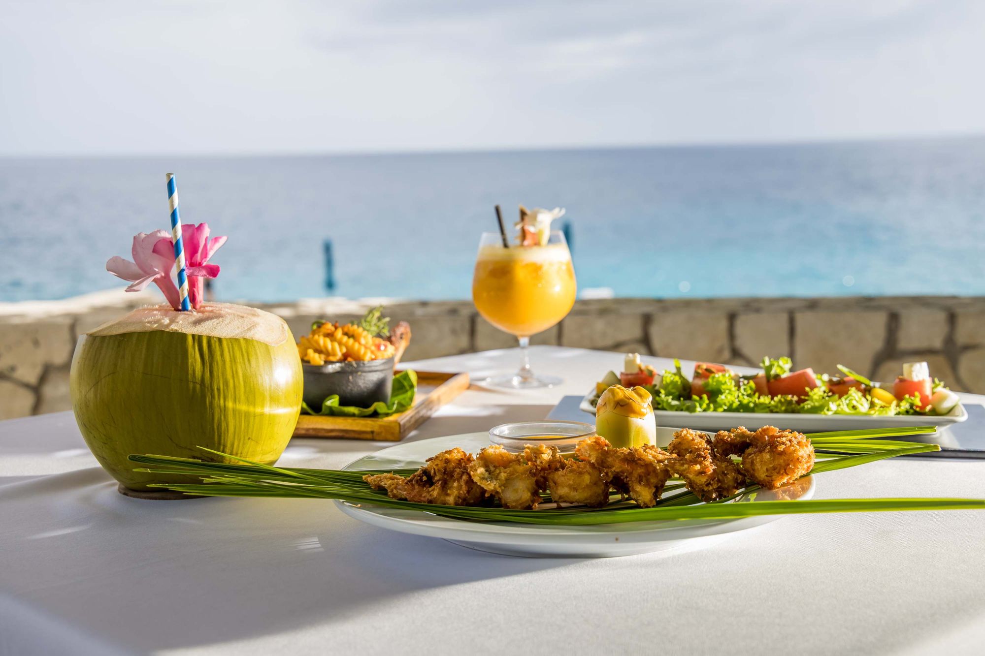 A beachside table set with a carved coconut drink, flowers, grilled skewers, and fresh salad with the ocean in view.