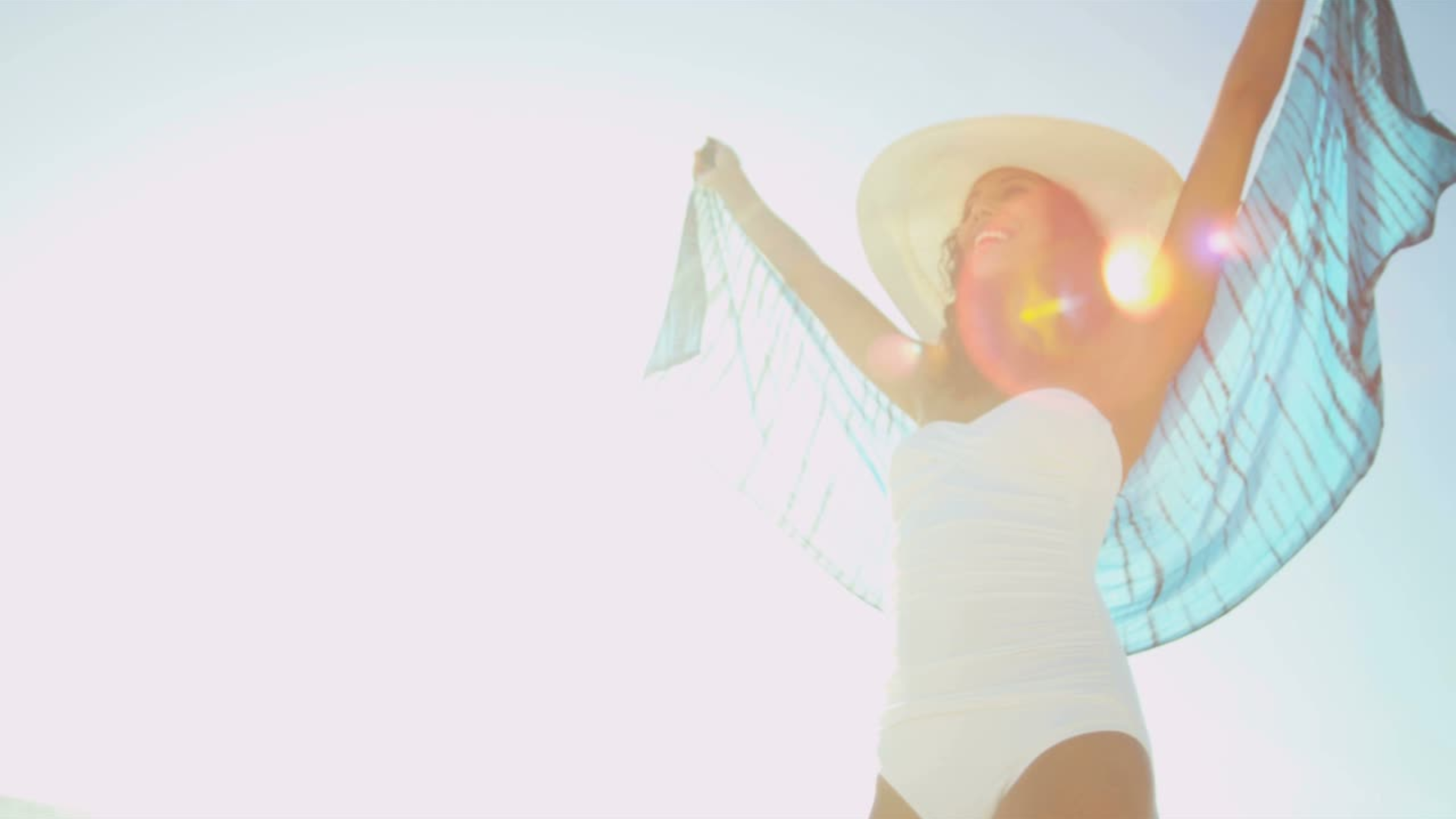 A woman in a white swimsuit stands under bright sun, wearing a wide-brim hat and a sheer blue cover, arms raised joyfully.
