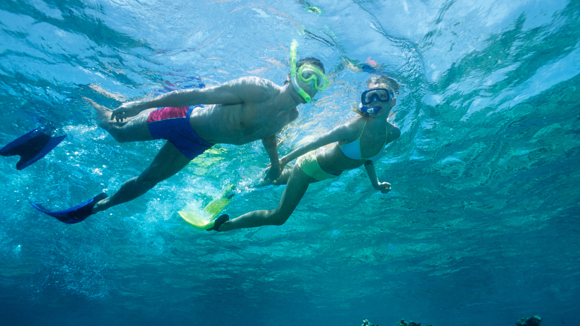 Two people snorkeling underwater, wearing masks and fins, swimming side by side near a bright blue seascape.