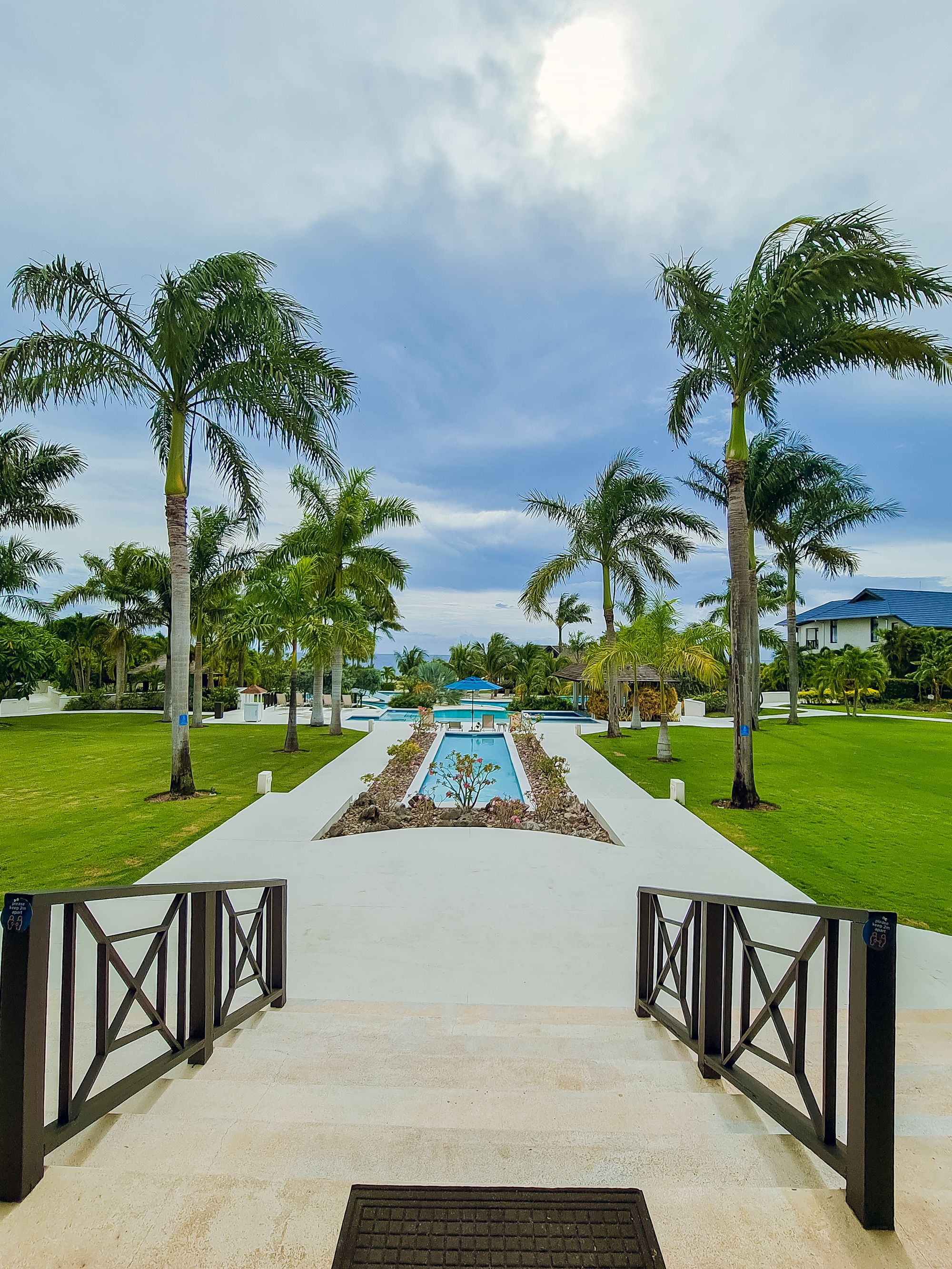 Palm trees line a manicured lawn with a central rectangular pool, a walkway, and a seaside view beyond the ocean horizon, sunny and relaxing.