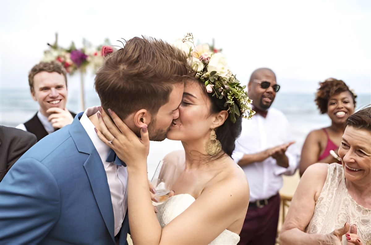 A newly married couple kiss at their outdoor beach wedding, friends and family cheering in the background.