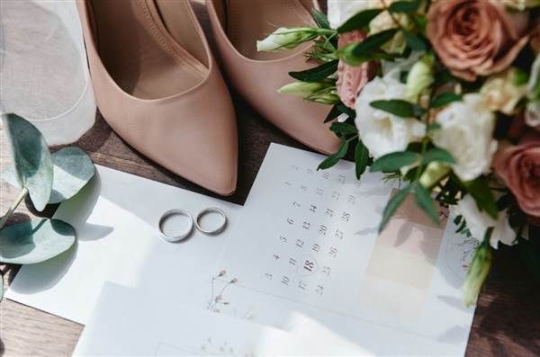 A wedding scene: blush heels, a bouquet with pink and white roses, rings, and a calendar spread on a table with soft light.