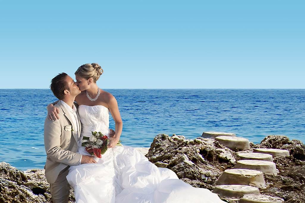A newlywed couple in wedding attire share a kiss by the rocky shoreline with the sea in the background, posing on a stone staircase.