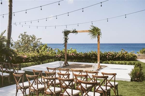 Outdoor wedding setup by the sea with string lights, wooden chairs, and a simple wooden arch over a sandy shore backdrop.