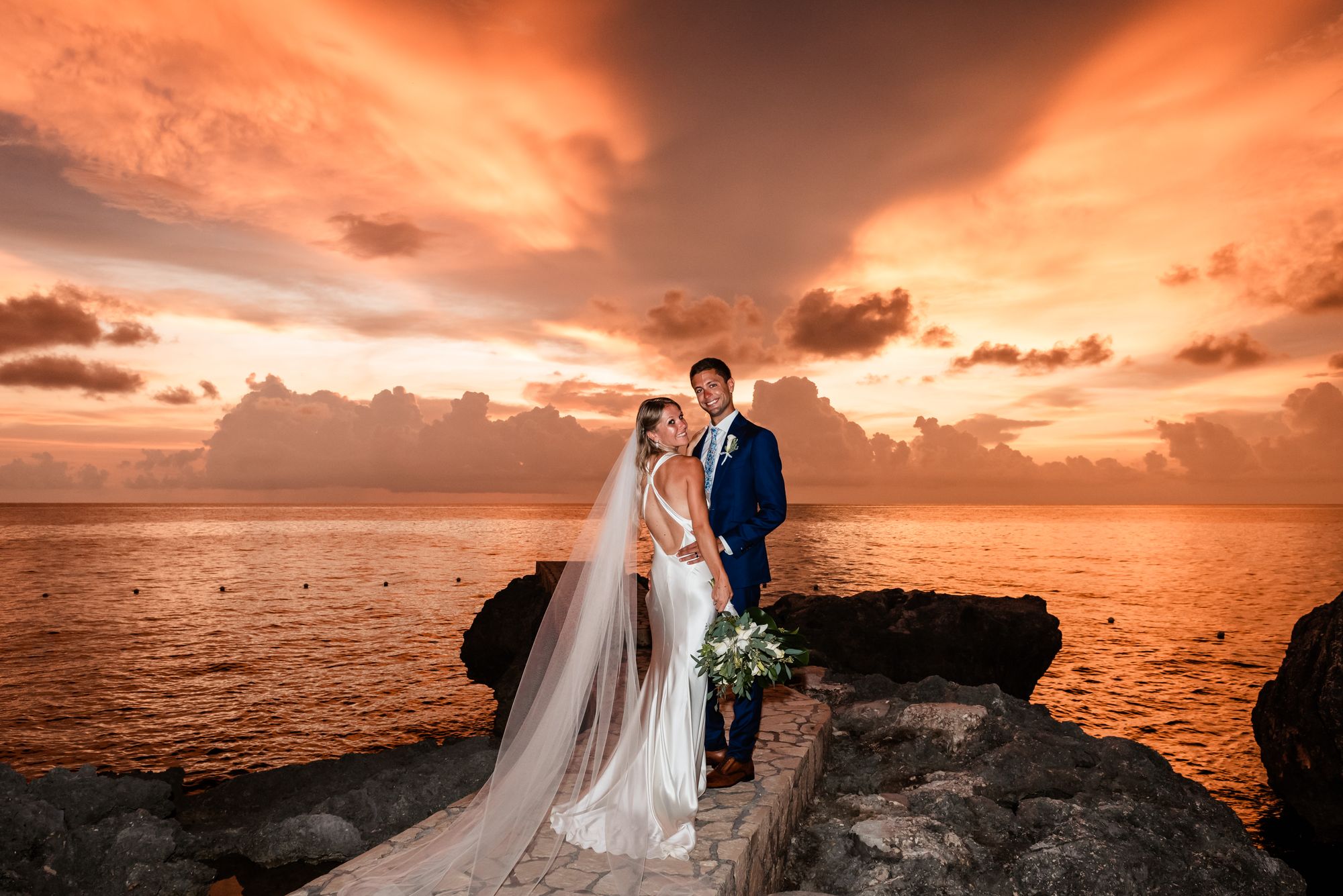 Wedding couple on a rocky seaside at sunset, with the ocean and dramatic clouds behind them, ready for a lifelong “yes.”