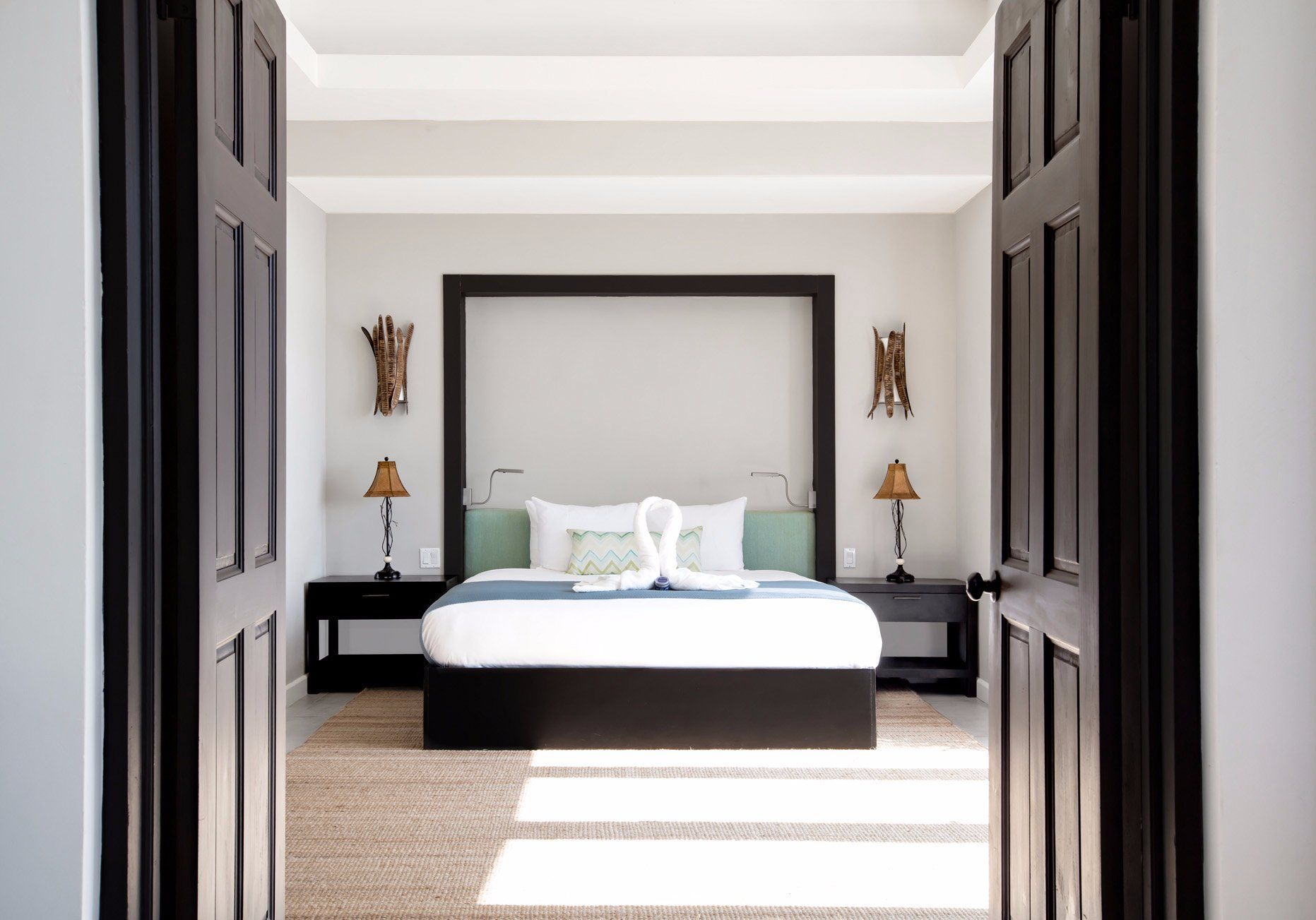 A bright, modern bedroom framed by dark double doors, with a large bed, two black nightstands, lamps, and neutral decor.
