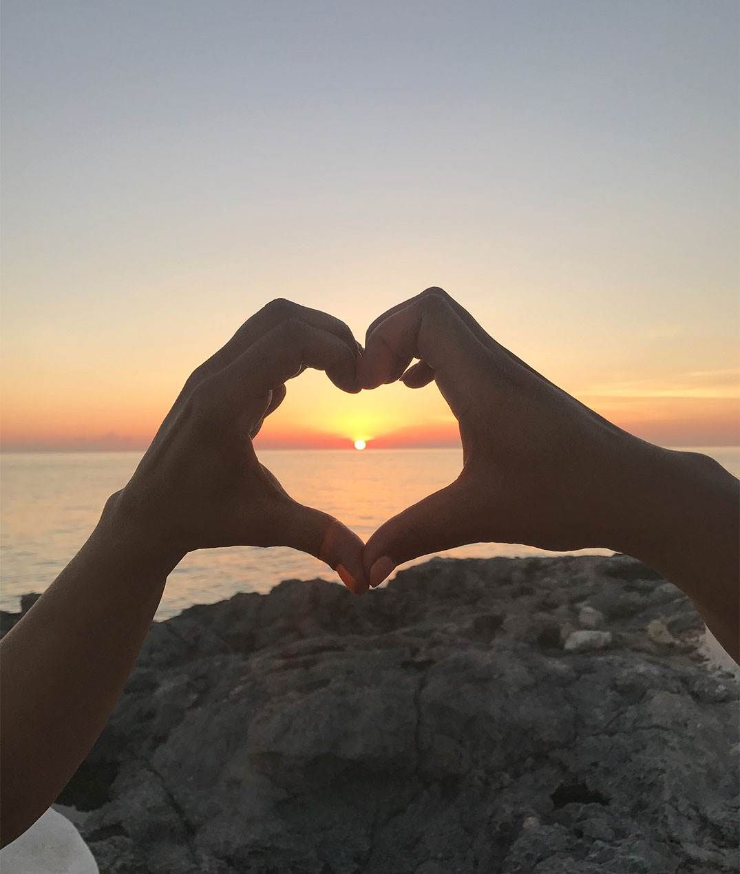 Two hands form a heart frame around a setting sun over the ocean, with a rocky shoreline in the foreground.