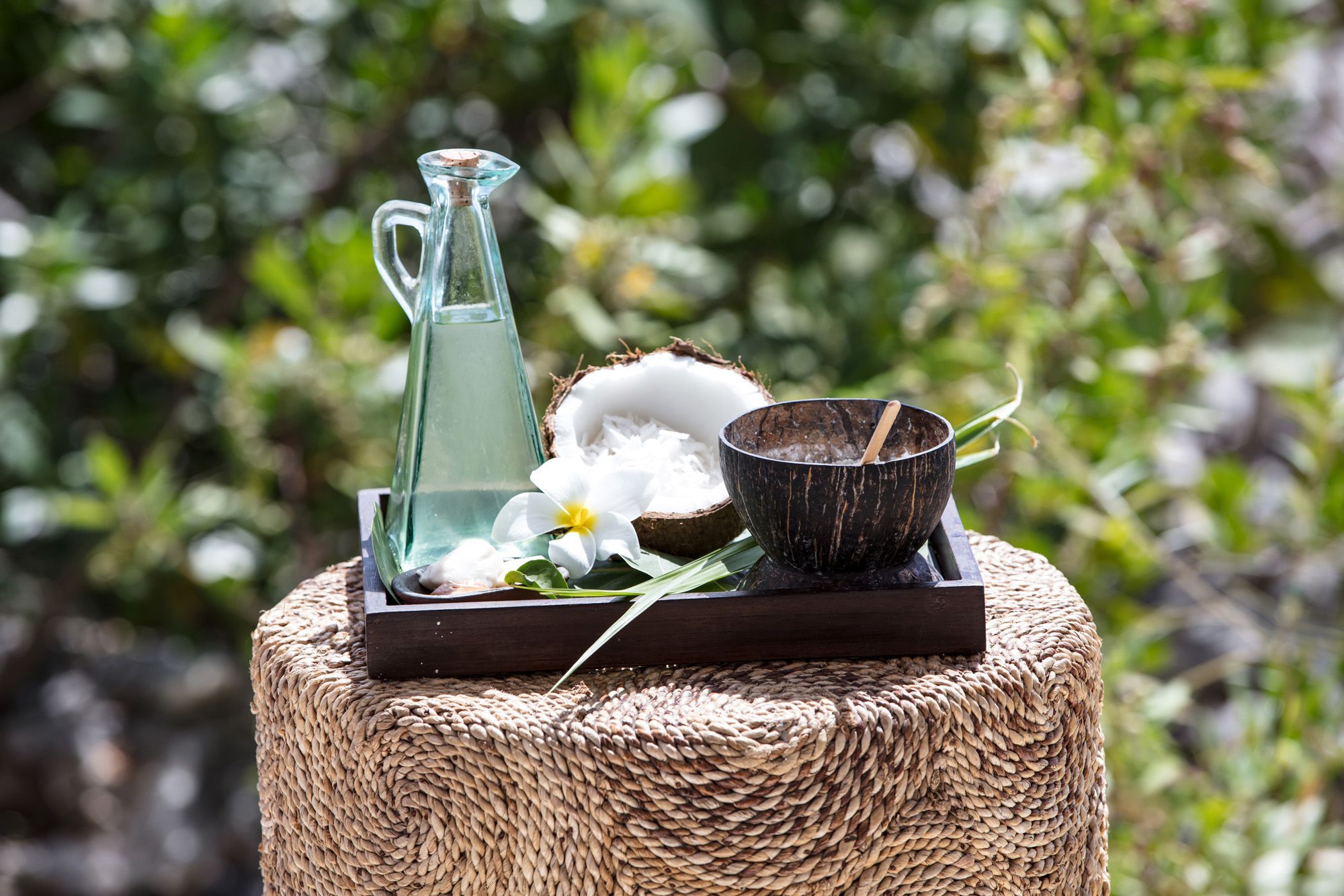 A glass bottle with a narrow neck on a wooden tray, plus a small wooden bowl and a coconut husk, placed on a woven stool outdoors, with greenery in the background.