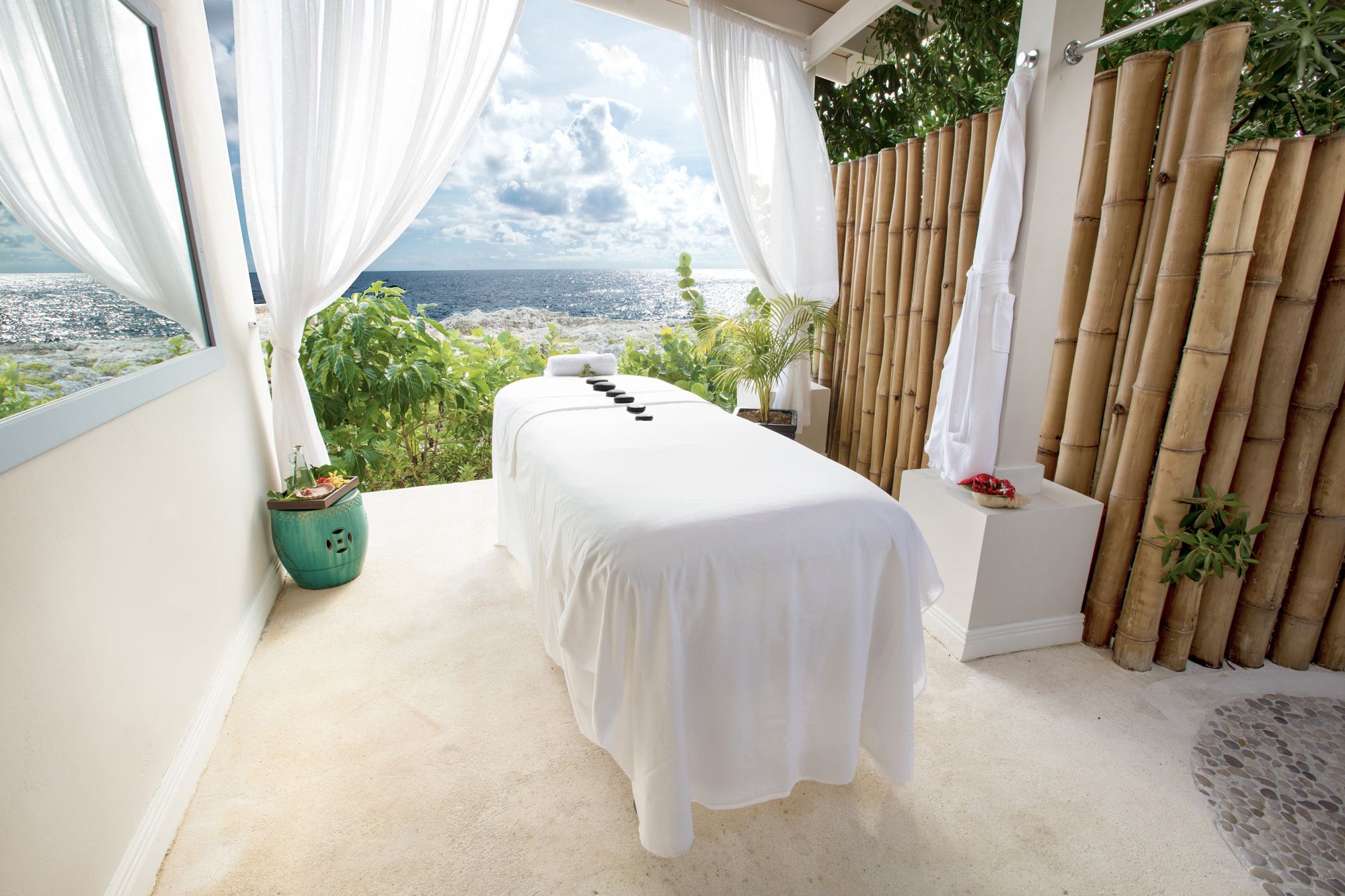 A seaside spa terrace with a white treatment bed, bamboo curtain walls, tropical plants, and an ocean view through open curtains.