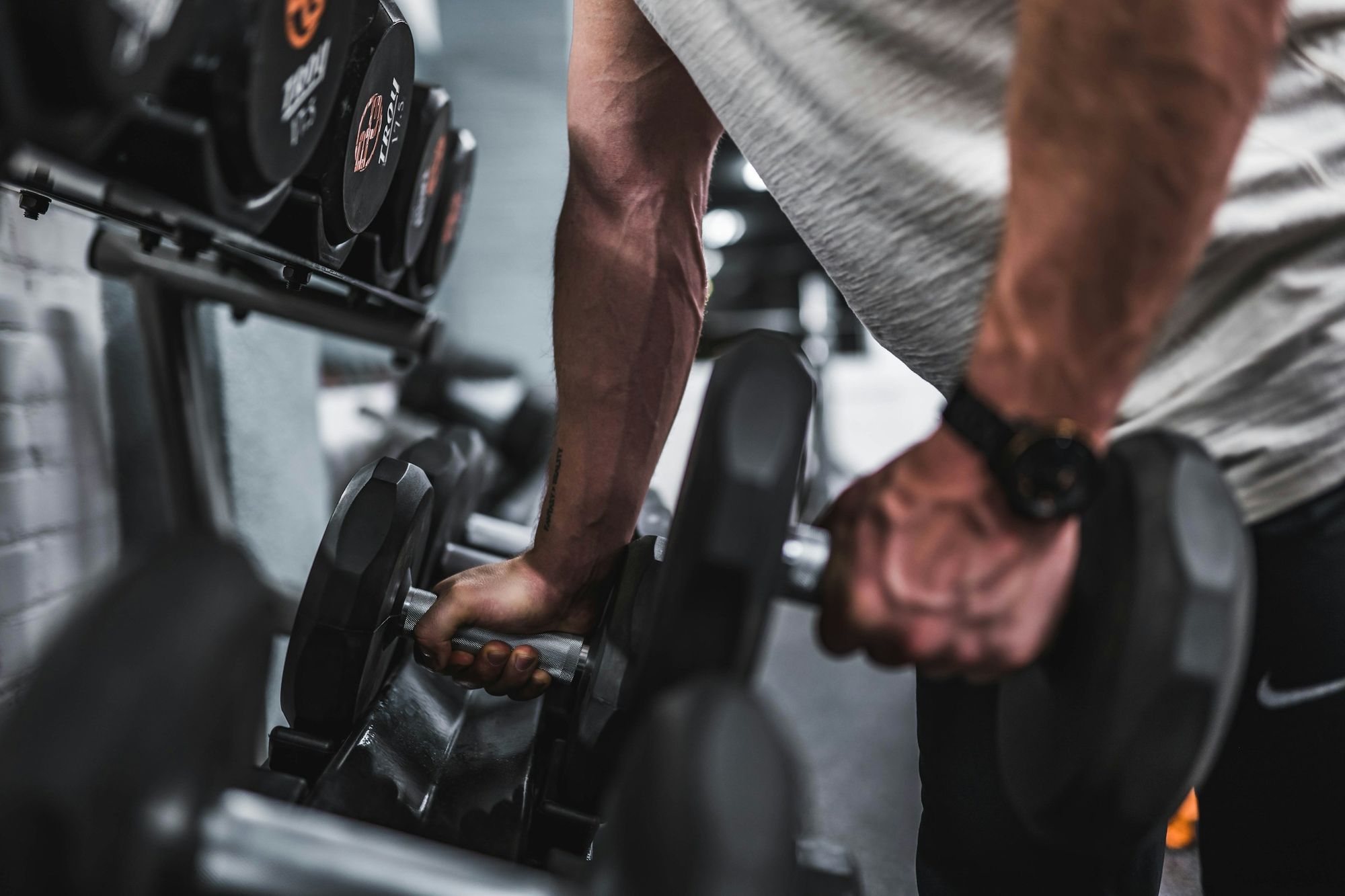 A person gripping a dumbbell near a rack of weights, mid-workout, showing focused effort and a gym setting.