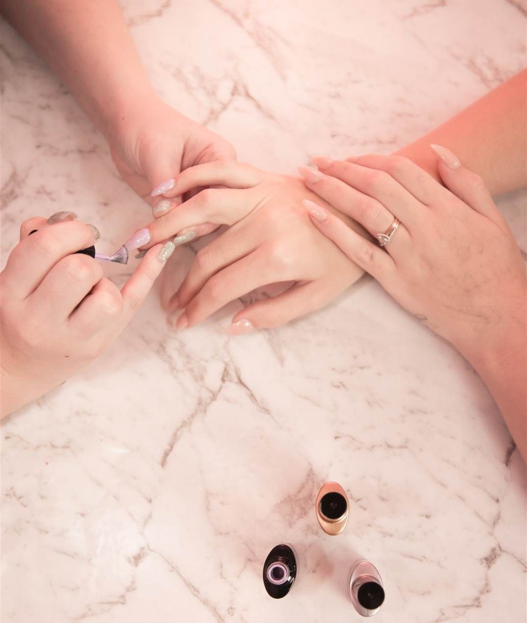 Two people getting a manicure, one hand receiving nail care while the other guides, with nail polish bottles on a marble surface.