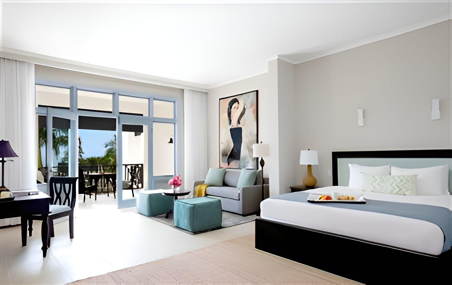 A bright hotel room with a king bed, seating area, and a balcony/patio. Modern decor, soft grays and whites, with a large window and framed art.