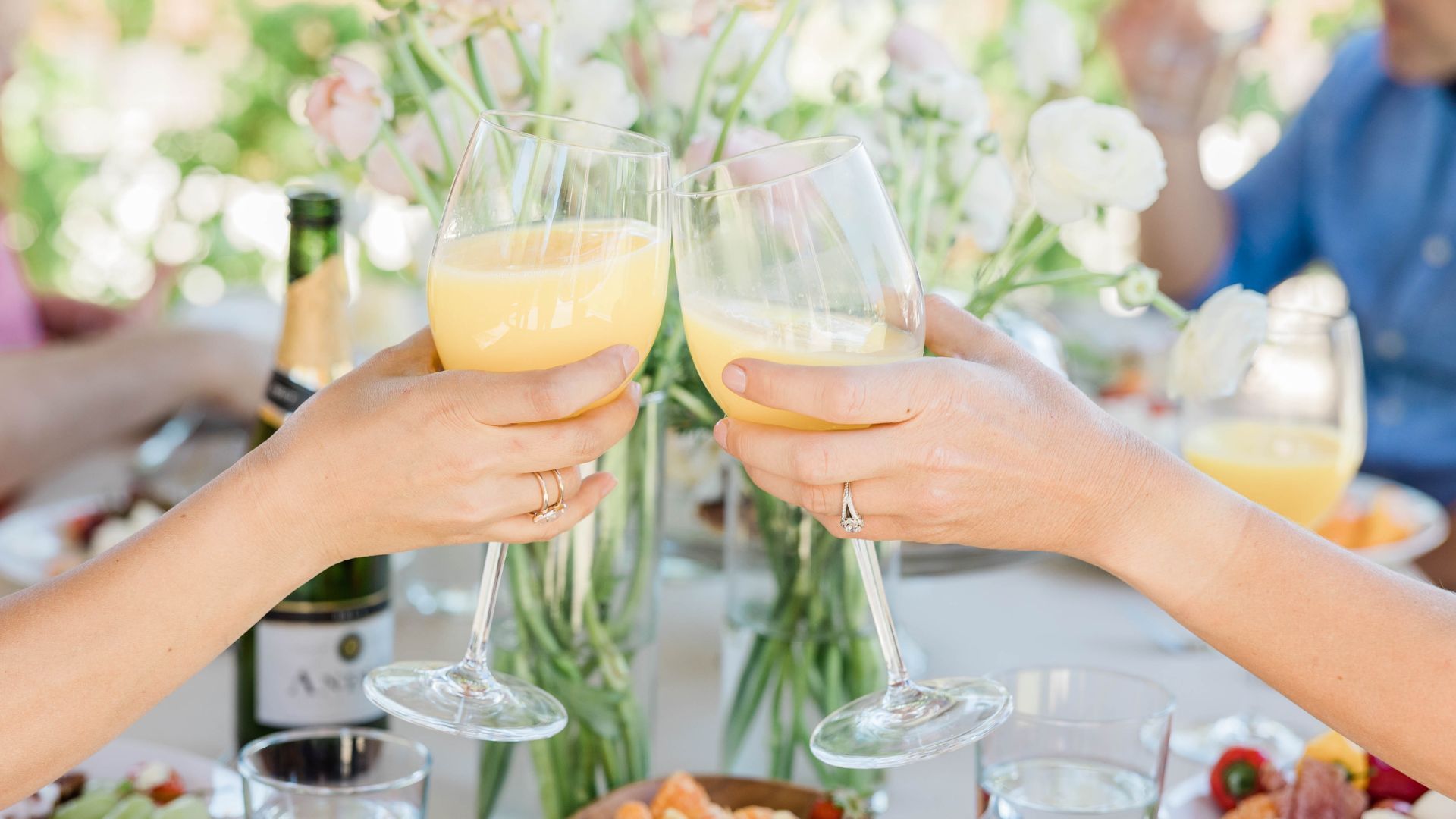 People clinking wine glasses in a cheerful toast at a table with flowers and drinks.