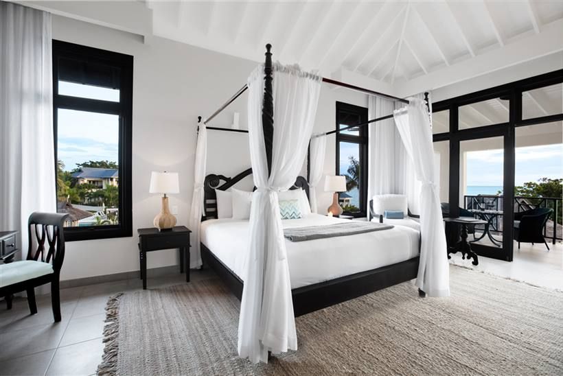 A luxurious white-canopy bed anchors a bright, airy bedroom with black-framed windows, a woven rug, and a balcony view—elegant coastal chic.