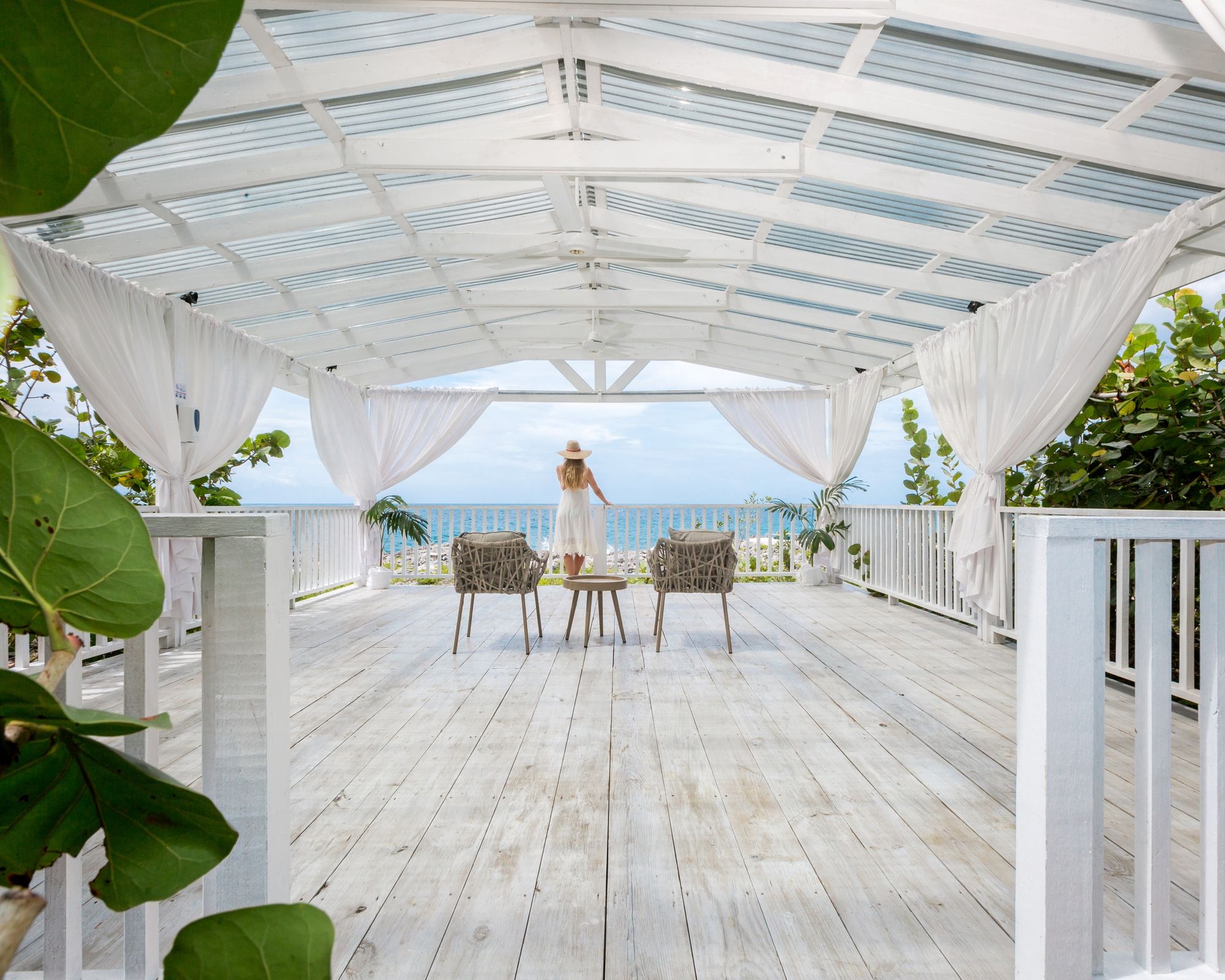 A person stands at a bright, airy white wooden deck pavilion overlooking the ocean, with chairs and a table set under draped curtains.