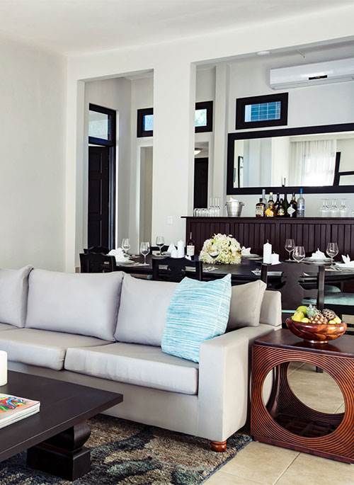 A modern living room with a white sofa, coffee table, windowed wall bar, and neatly arranged drinks and flowers.