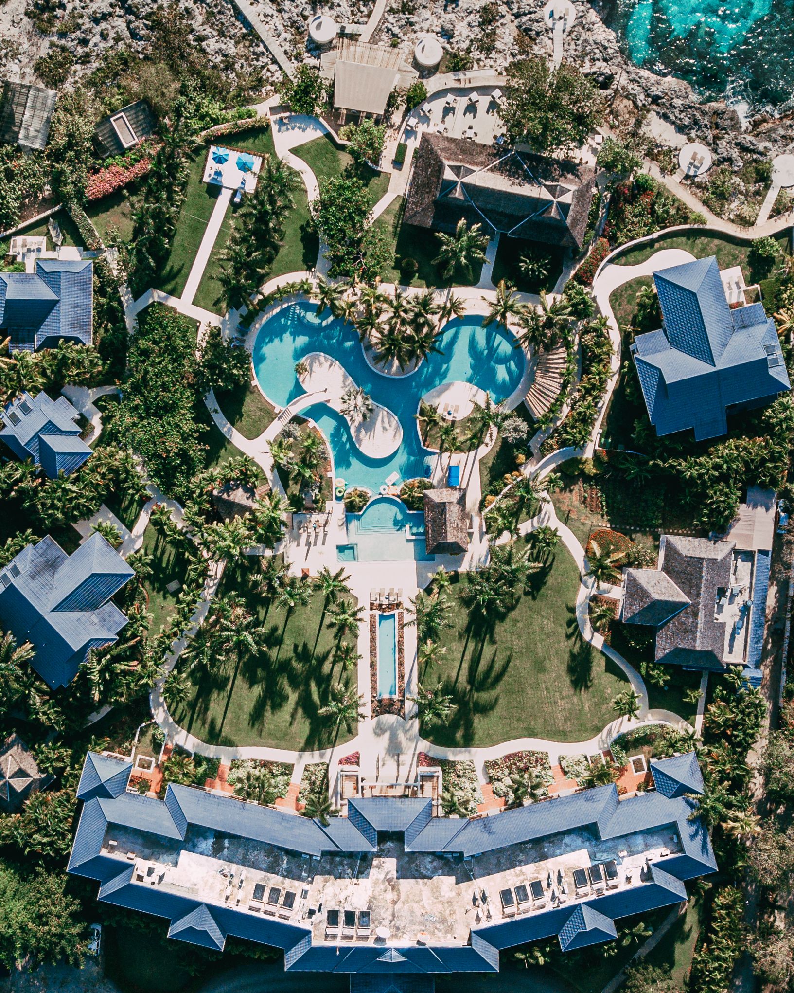 An aerial view of a tropical resort with blue pools, sun loungers, and lush gardens surrounding villas and cabanas.