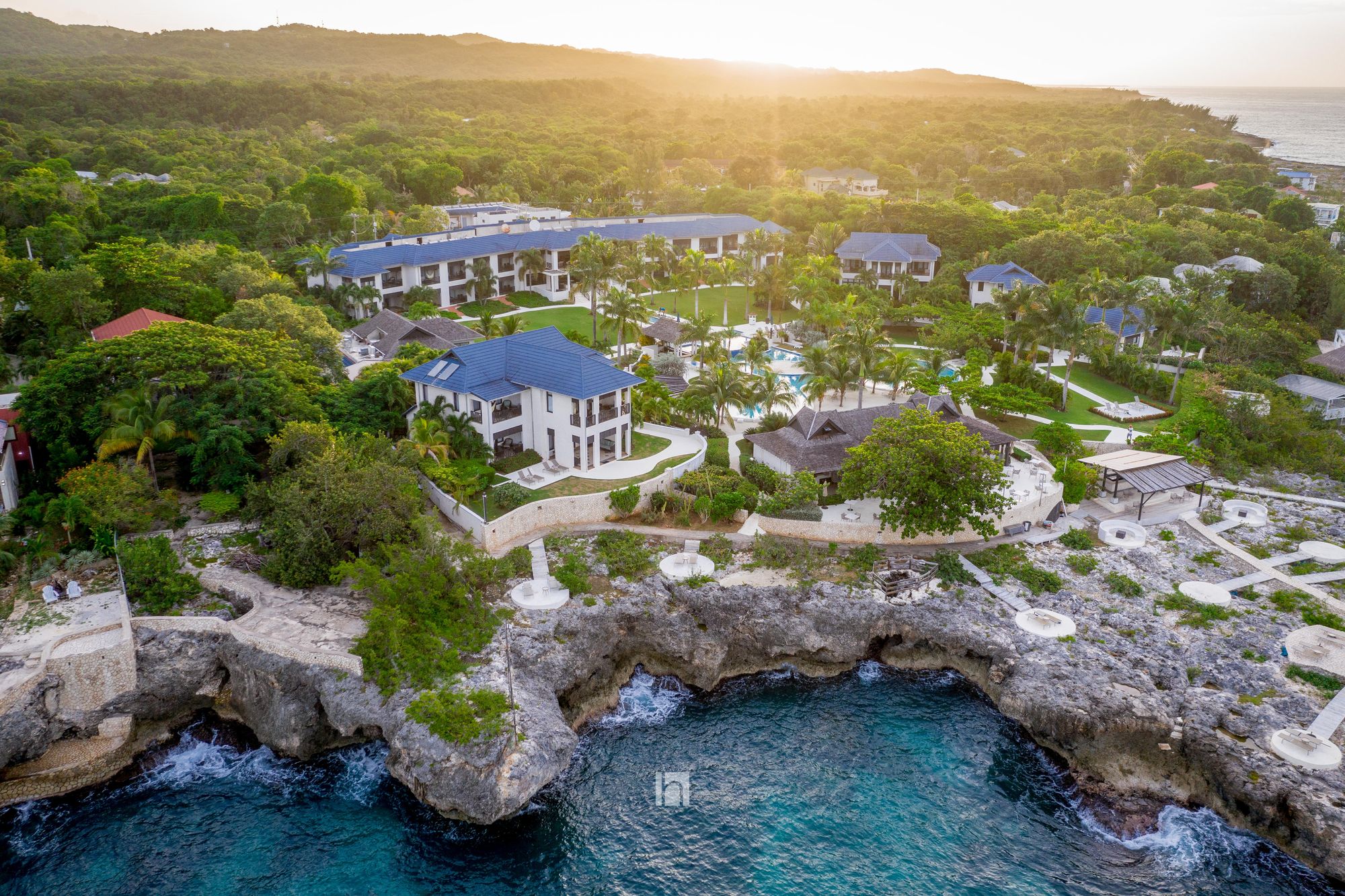Aerial view of a tropical beachfront resort with blue-roof villas, lush palms, and a rocky cove at sunset.