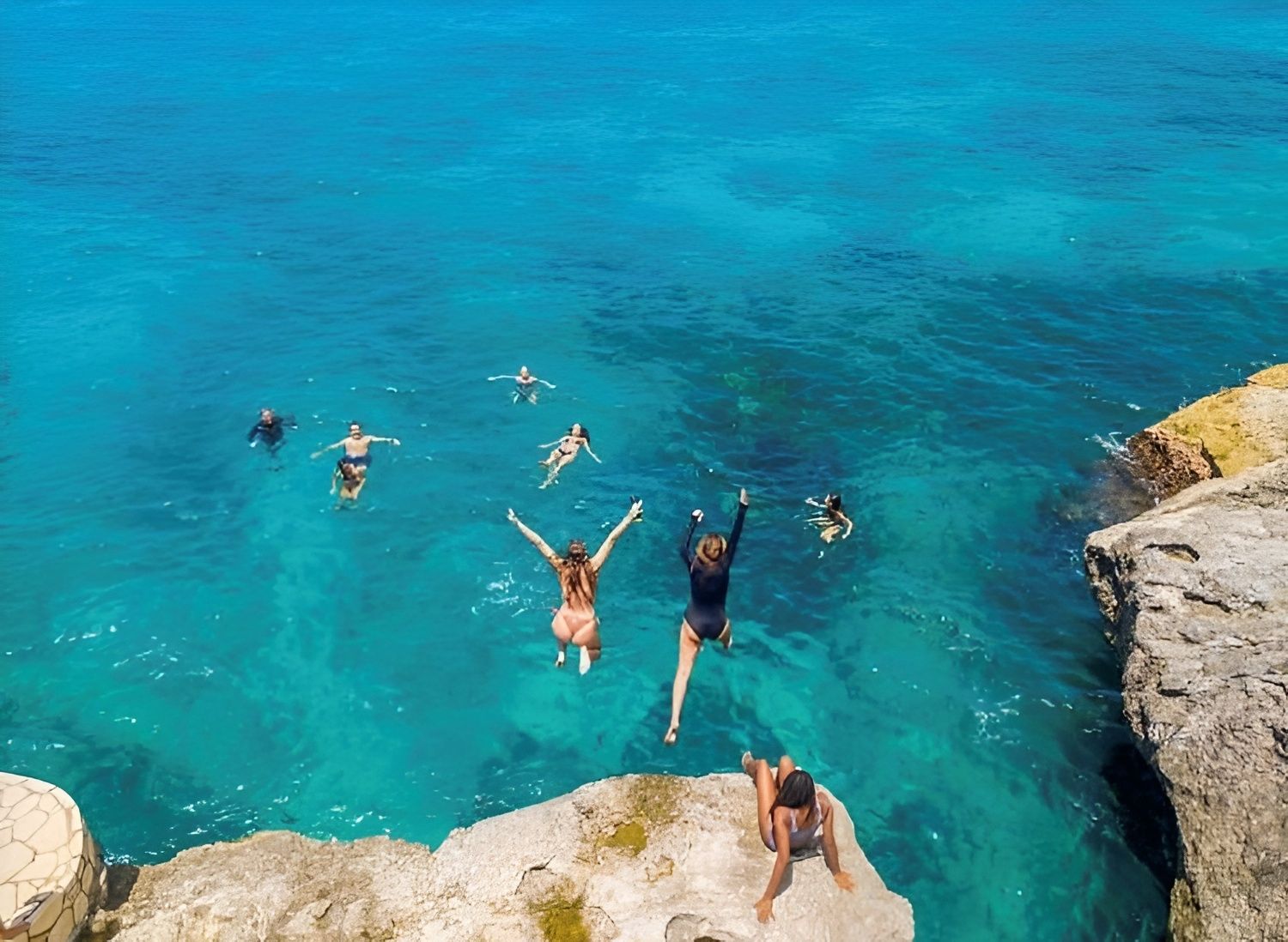 People jump into a clear turquoise ocean from a rocky cliff, swimming and diving in open water.