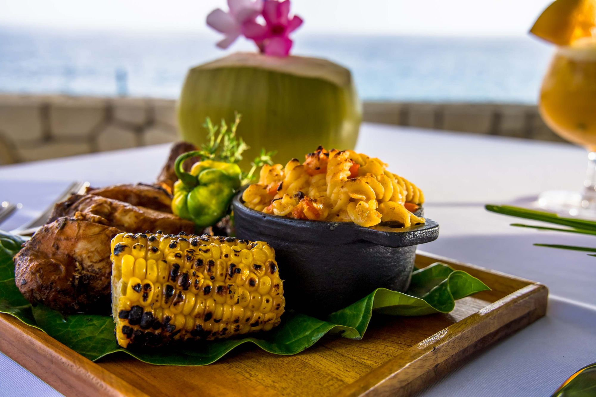 A tropical seafood dish with grilled corn, roasted plantain, and a saffron-tleck rice or side, served on a wooden board with a coconut drink, by the sea.
