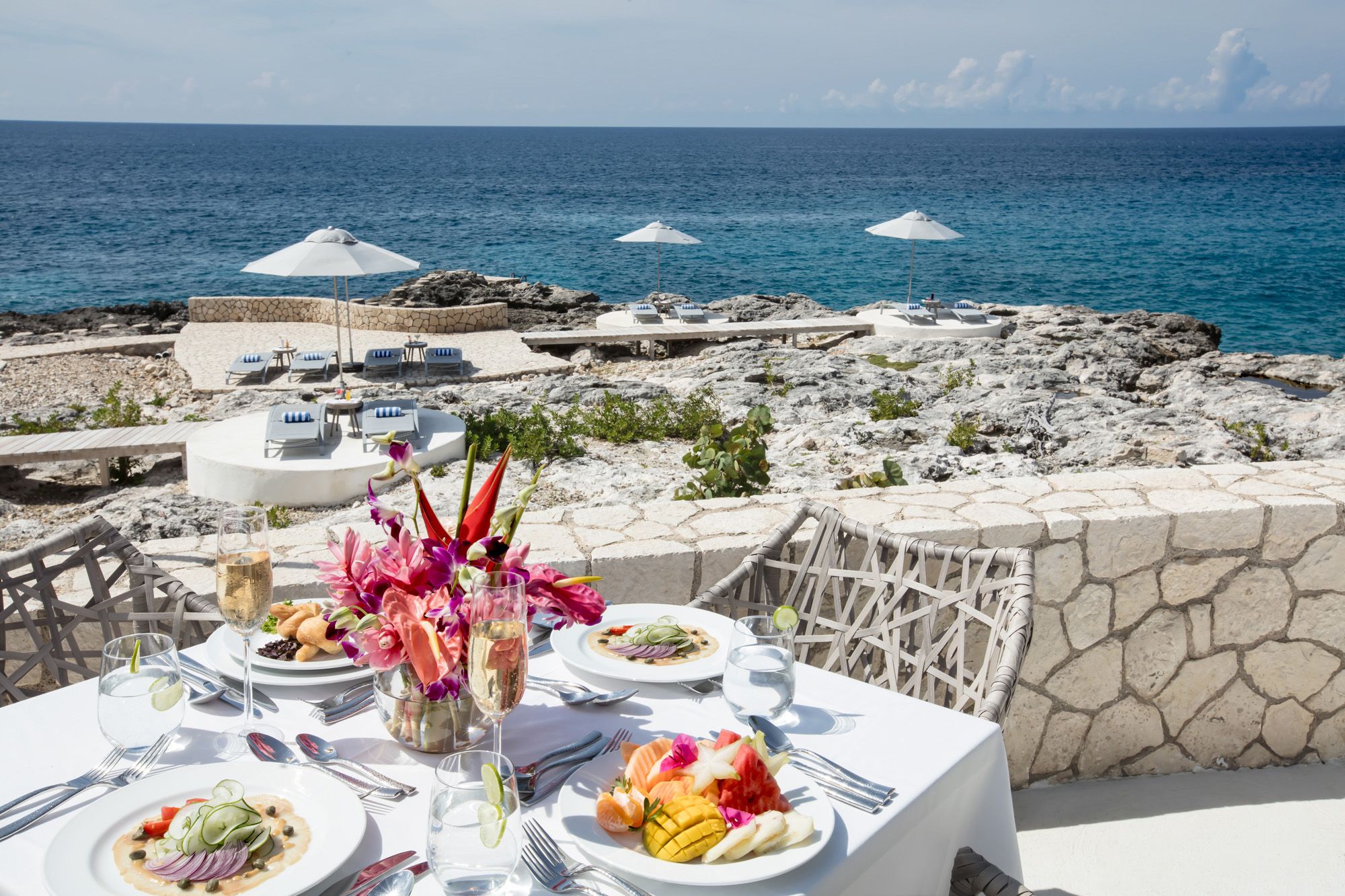 A seaside restaurant table set with white tablecloths, plates of food, flowers, and a view of the rocky shore and blue sea under a sunny sky.