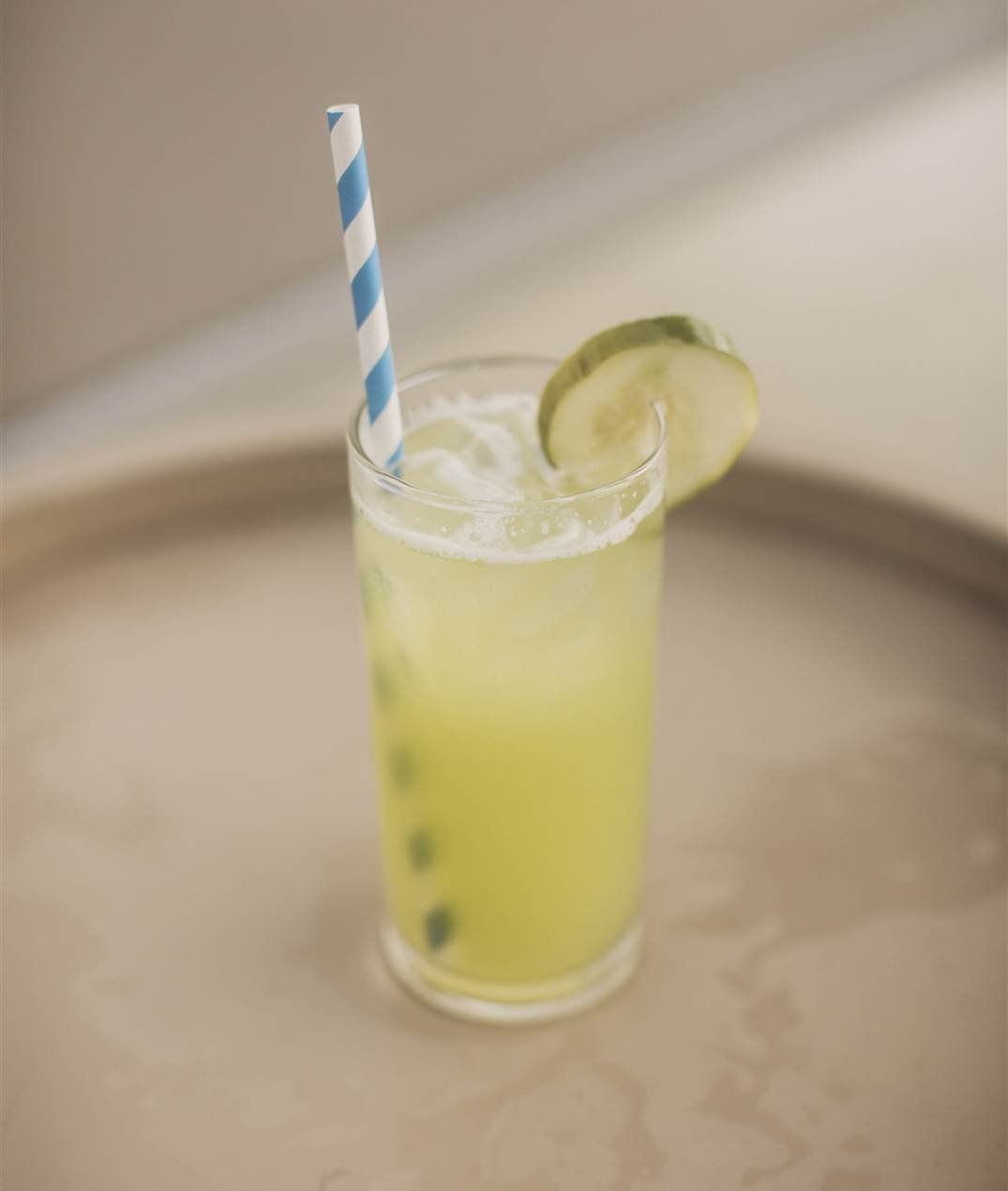 A tall glass of yellow lime drink with a striped blue straw and a lime wedge garnish, on a metal tray, looks refreshing.