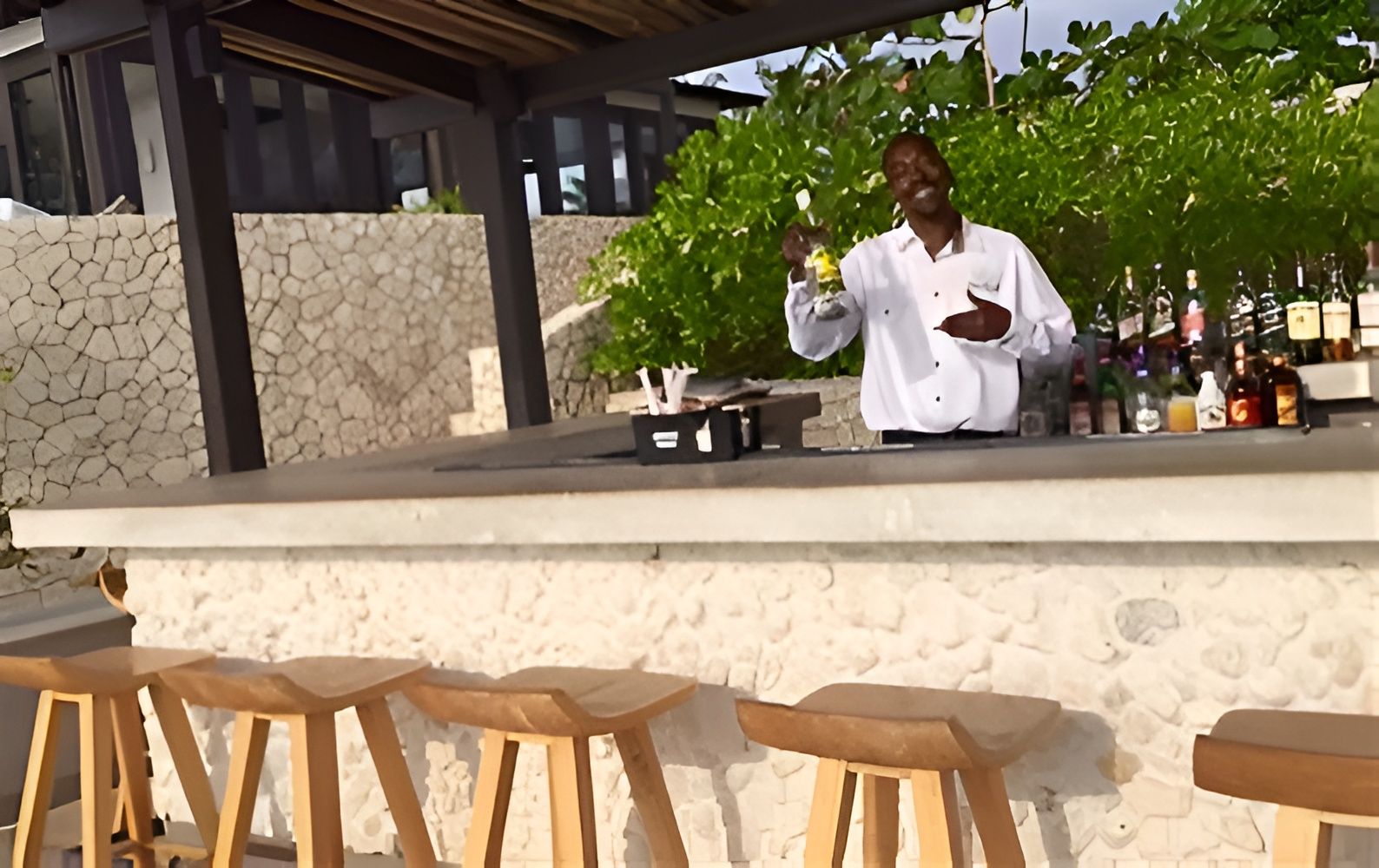 A bartender stands behind a white counter with bottles, chalkboard items, and wooden bar stools in an outdoor setting.
