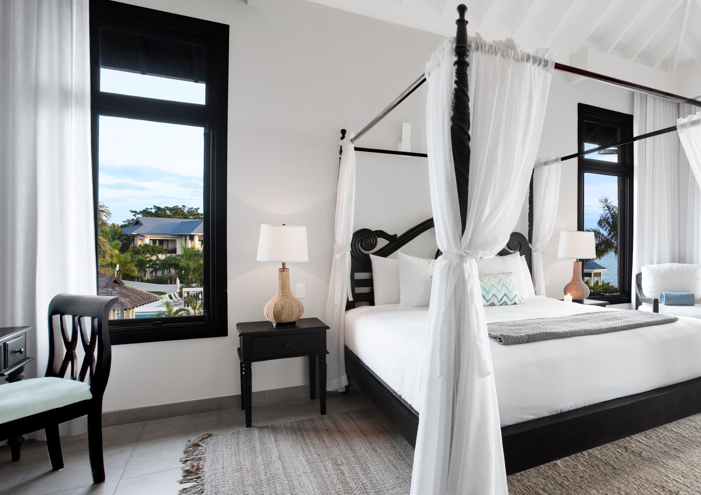 A bright, modern hotel room with a four-poster bed, white linens, black furniture, and large windows letting in natural light.