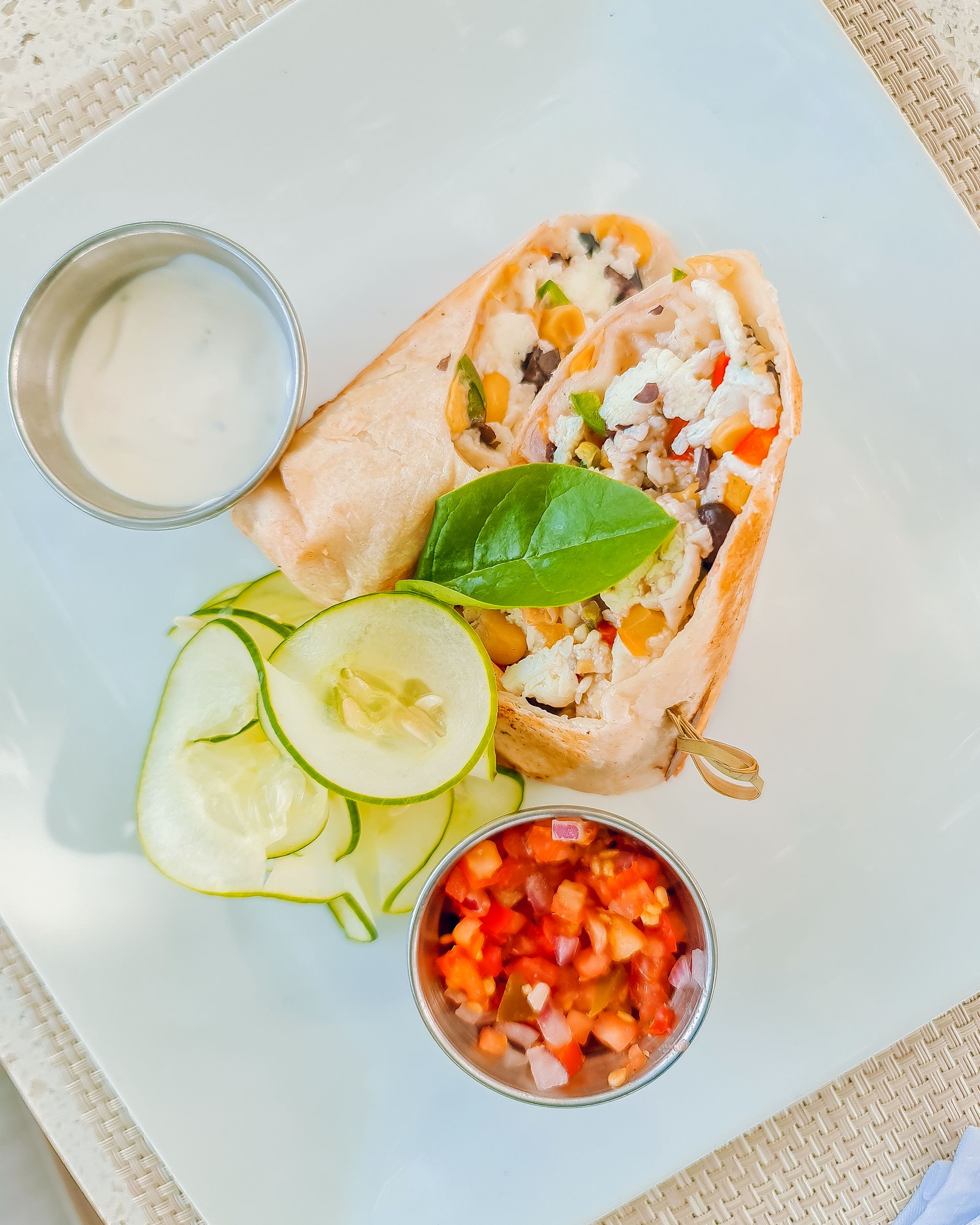 A veggie burrito on a plate with sliced cucumbers, a lime slice, a small bowl of salsa, and a dipping sauce on the side, delicious.