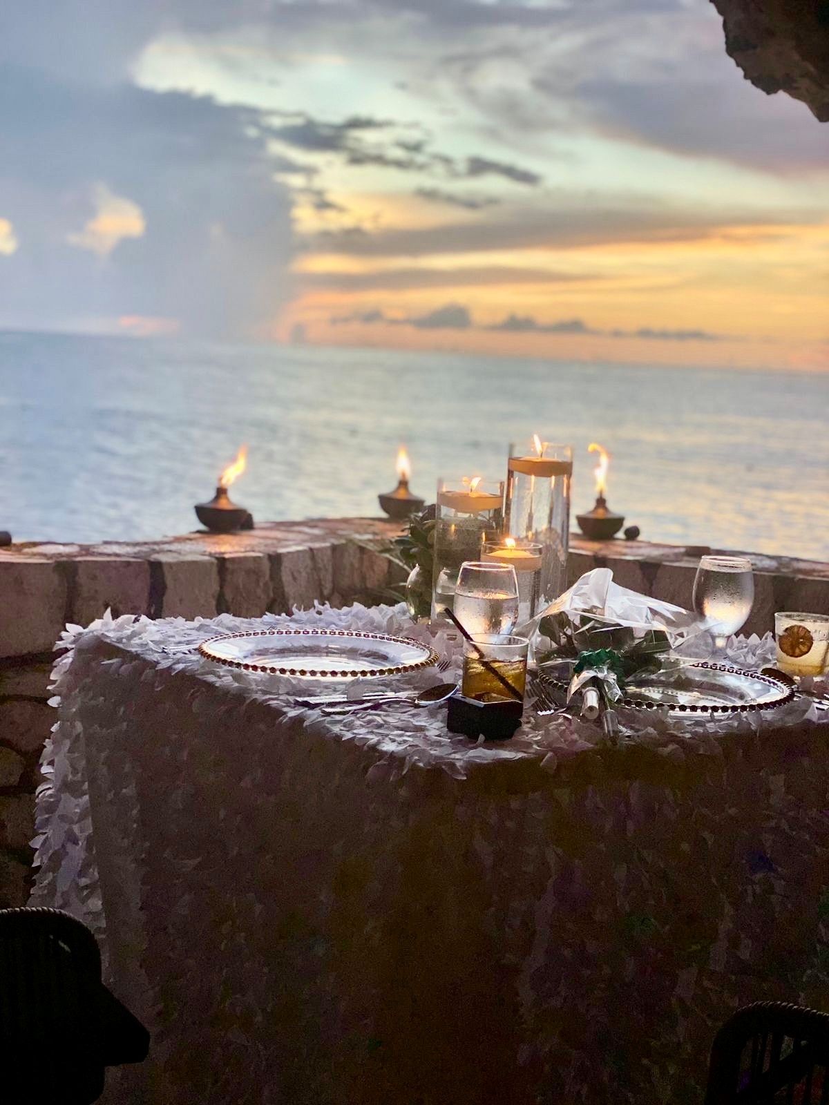 A romantic beachside dinner setup with candles, a lace tablecloth, and glowing sunset over the water, creating a serene, intimate scene.