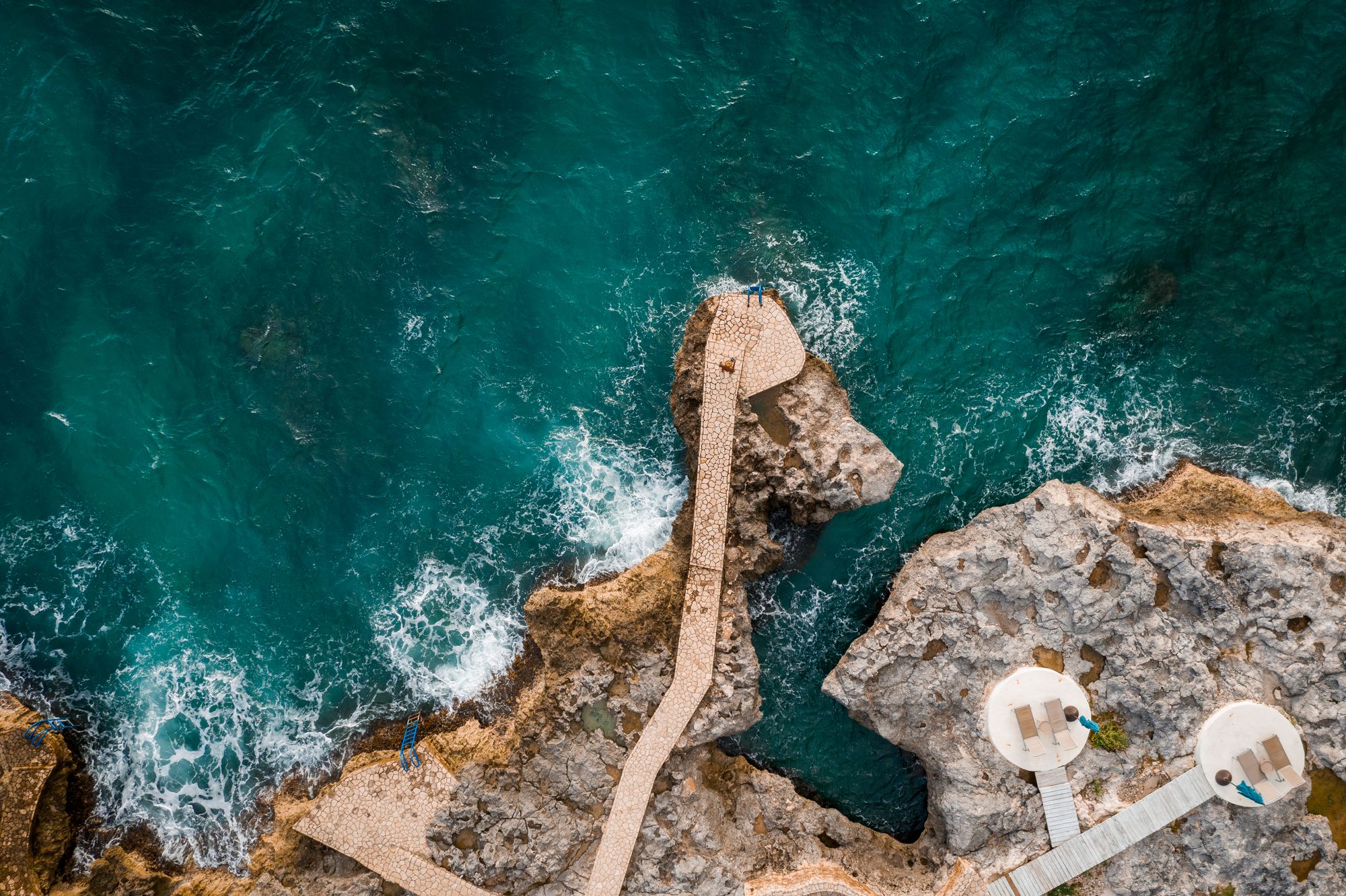 Aerial view of rugged rocky coastline with a concrete jetty extending into turquoise water, small white circular platforms at the edge, waves crashing below.