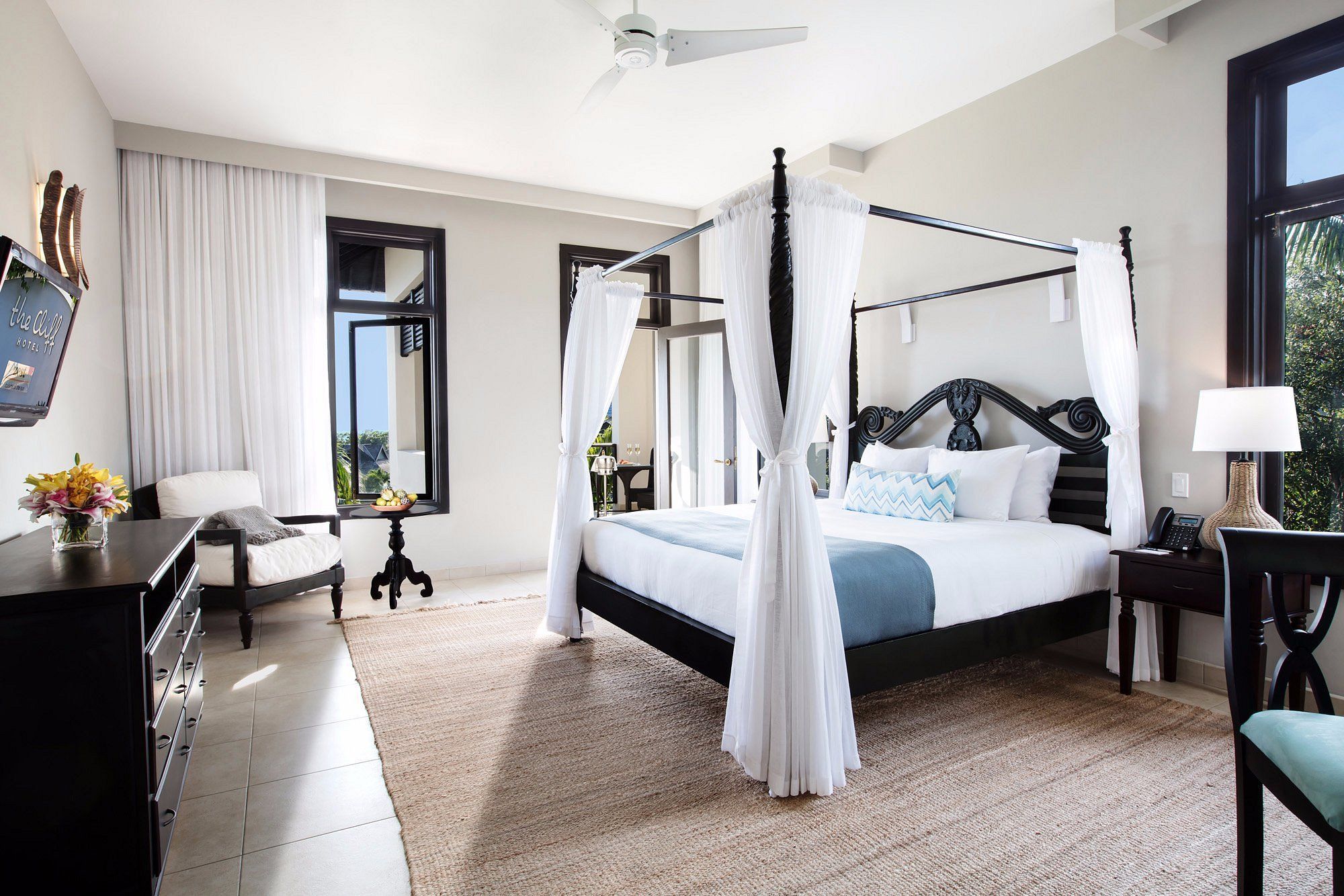 A luxurious four-poster bed with white drapes sits in a bright hotel room, flanked by nightstands, a desk, and seating by windows.