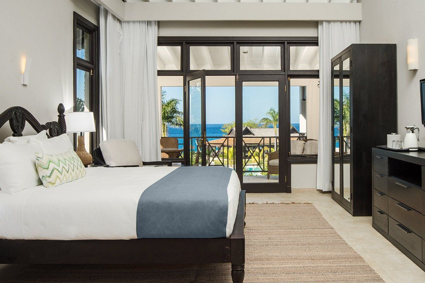A hotel room with a dark wooden bed, white and blue bedding, a rug, and a balcony view of palm trees and the ocean through glass doors. Ending with a period.