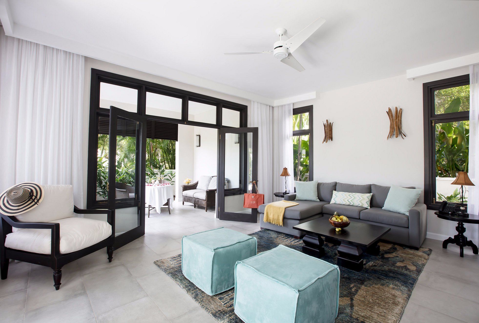 A bright, airy living room with a gray sofa, two turquoise ottomans, a glass coffee table, and large windows opening to a patio, sunlit and stylish.