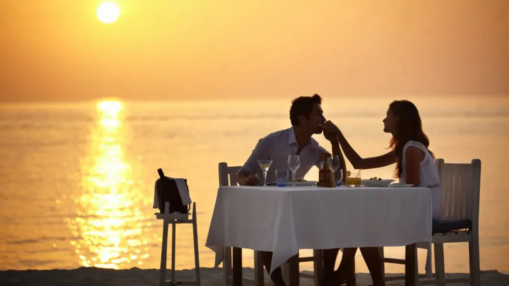A couple at a table for two by the sea at sunset, the sun low over the water.