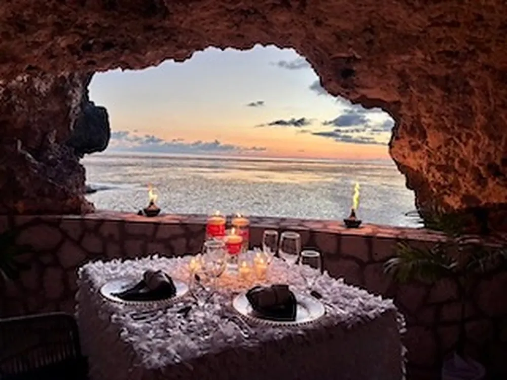 A table for two set with candles and rose petals inside The Cave, framed by the cave mouth at sunset over the sea.